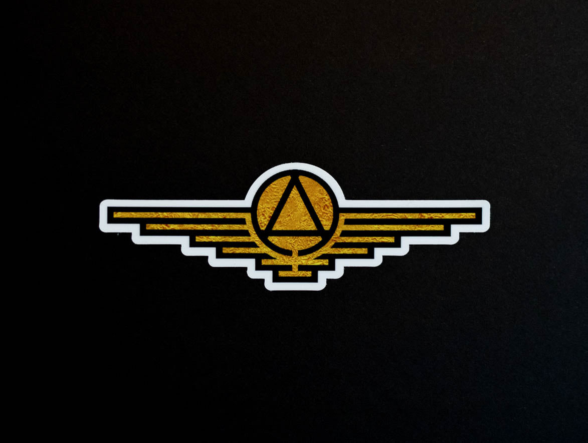 Gold sticker with black outlines depicting a triangle enclosed in a circle with wings extending on either side