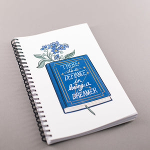 Spiral notebook with white background, blue flowers in a book that says "there is a defiance in being a dreamer"