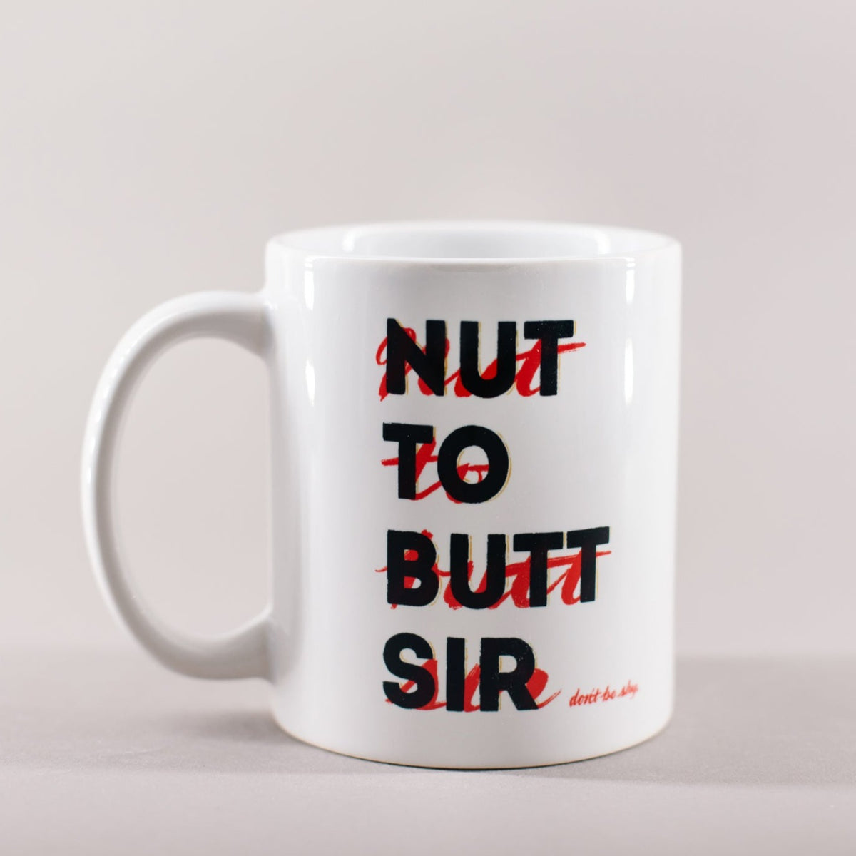White mug with black and red whiting that says 'Nut to butt sir, don't be shy'