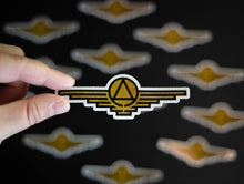 Load image into Gallery viewer, Gold sticker with black outlines depicting a triangle enclosed in a circle with wings extending on either side
