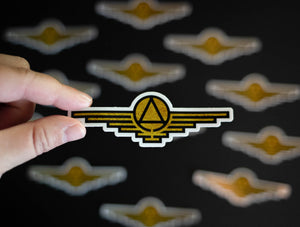 Gold sticker with black outlines depicting a triangle enclosed in a circle with wings extending on either side