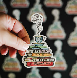 Sticker depicting a steaming tea cup and saucer on top of a stack of books that say "Books, she has found, are a way to live a thousand lives."