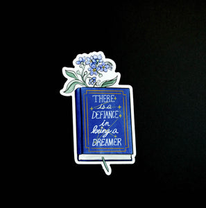 Sticker with blue flowers inside a hardcover blue book with "there is defiance in being a dreamer" and a constellation with seven stars 