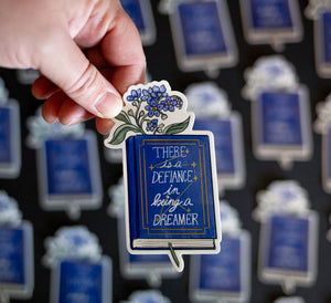 Sticker with blue flowers inside a hardcover blue book with "there is defiance in being a dreamer" and a constellation with seven stars on the cover