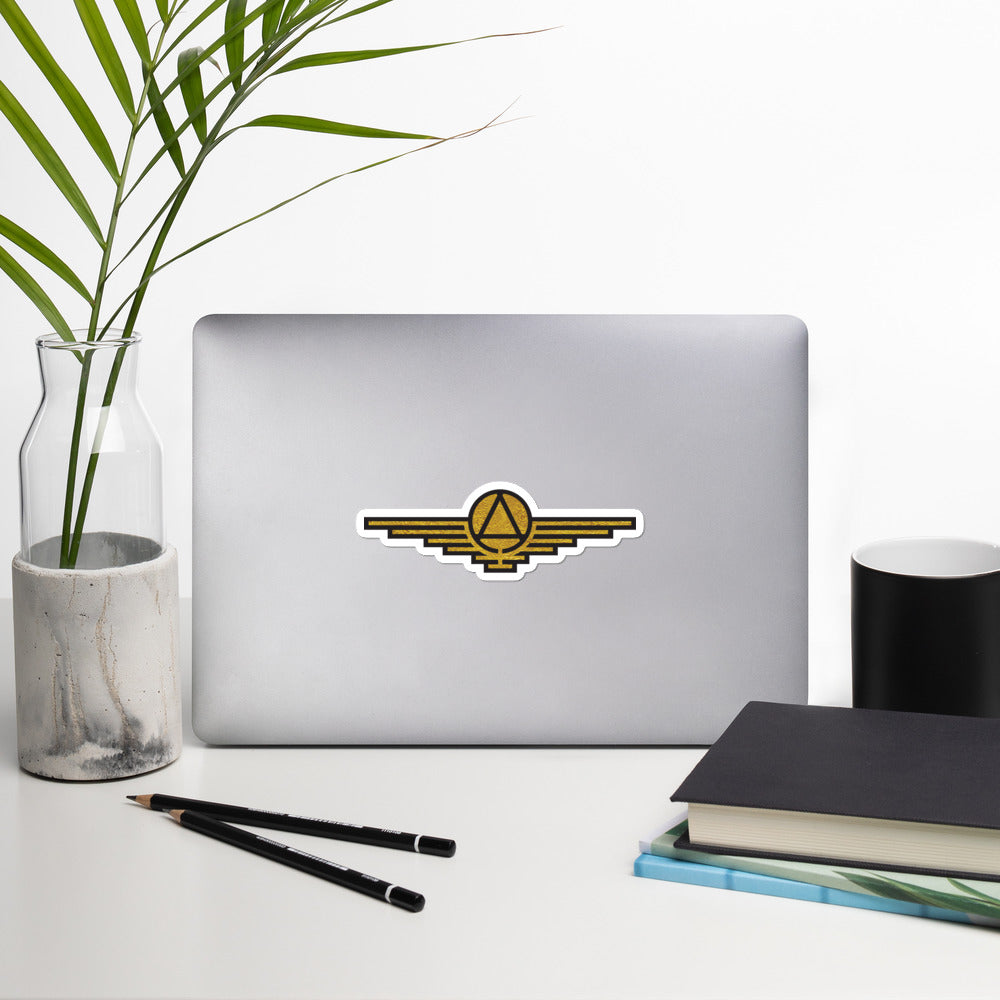 Gold sticker with black outlines depicting a triangle enclosed in a circle with wings extending on either side