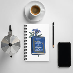 Spiral notebook with white background, blue flowers in a book that says "there is a defiance in being a dreamer"