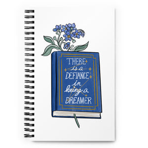 Spiral notebook with white background, blue flowers in a book that says "there is a defiance in being a dreamer"